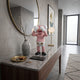 Petal Pink Iconify Jolly Figure, 30cm, covering eyes and laughing on a marble entryway console beside a round mirror and ceramic vase.