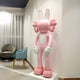 In a softly lit modern room, a Giant Sculptures 30cm Petal Pink Iconify Rabbit Figure with bunny ears, big gloves, blank face, and oversized feet stands near light blue and gray chairs by sheer white curtains. Crafted from resin.