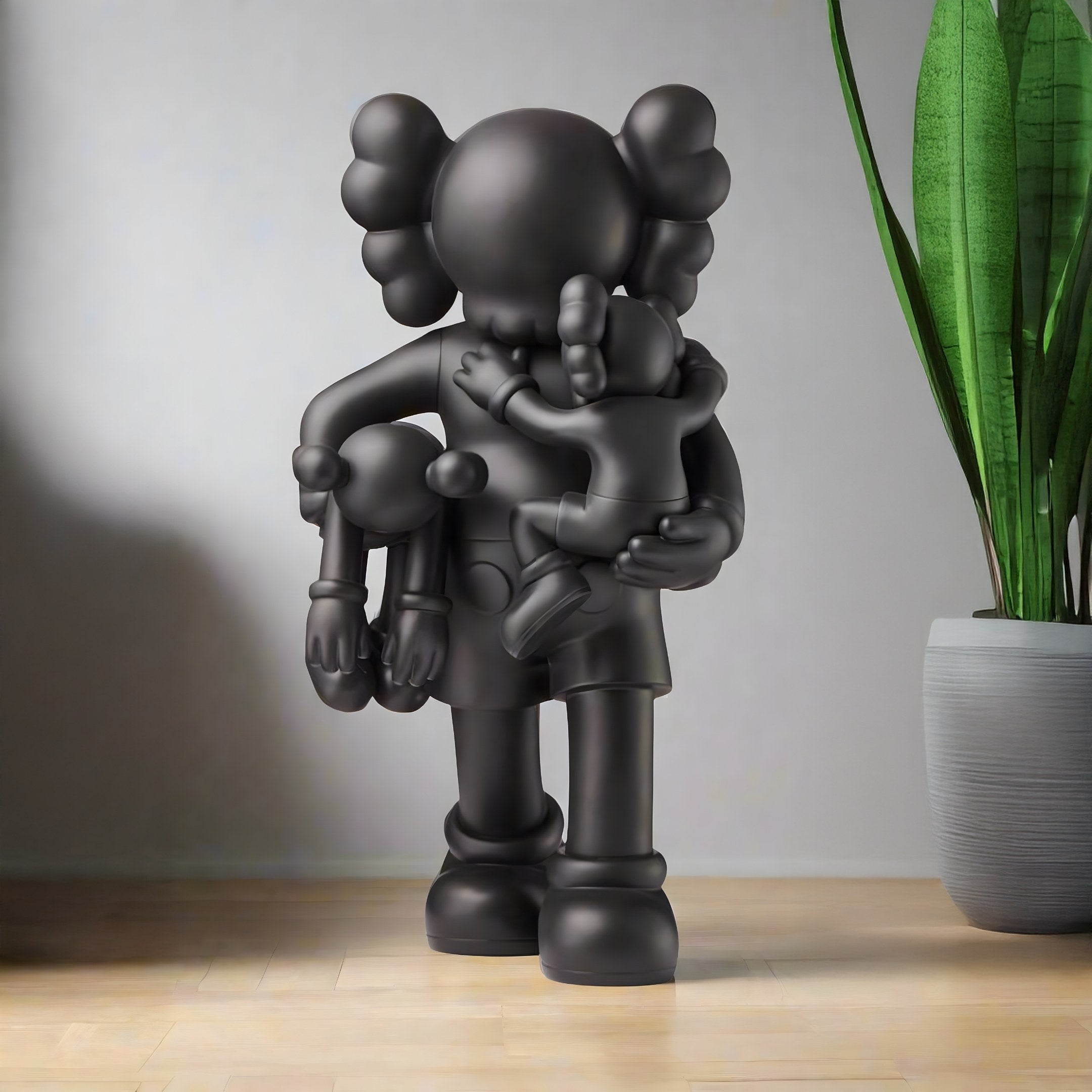 A 38cm Midnight Black Iconify Family Figure by Giant Sculptures stands on the wooden floor, featuring resin material, round ears, and simplified details while holding two smaller figures. A tall green potted plant nearby adds a modern touch against the plain wall.