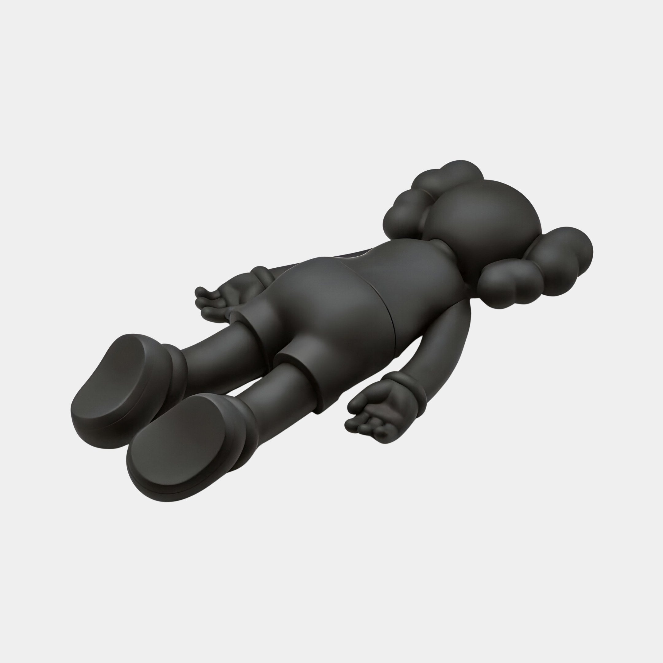 The Obsidian Black Iconify Flat Figure by Giant Sculptures is a 38cm cartoonish figure with bulbous limbs and a rounded head, lying flat on its back. Featuring exaggerated feet and mitten-like hands with a smooth glossy finish, it stands out beautifully against the plain light background.