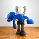 The Royal Blue Iconify Rescuing Figure by Giant Sculptures, 38cm tall, depicts a simplified figure holding a limp, furry blue character with a skull-like head and crossed eyes. It stands amidst modern interiors on a wooden floor with large, round ears in a graceful pose.