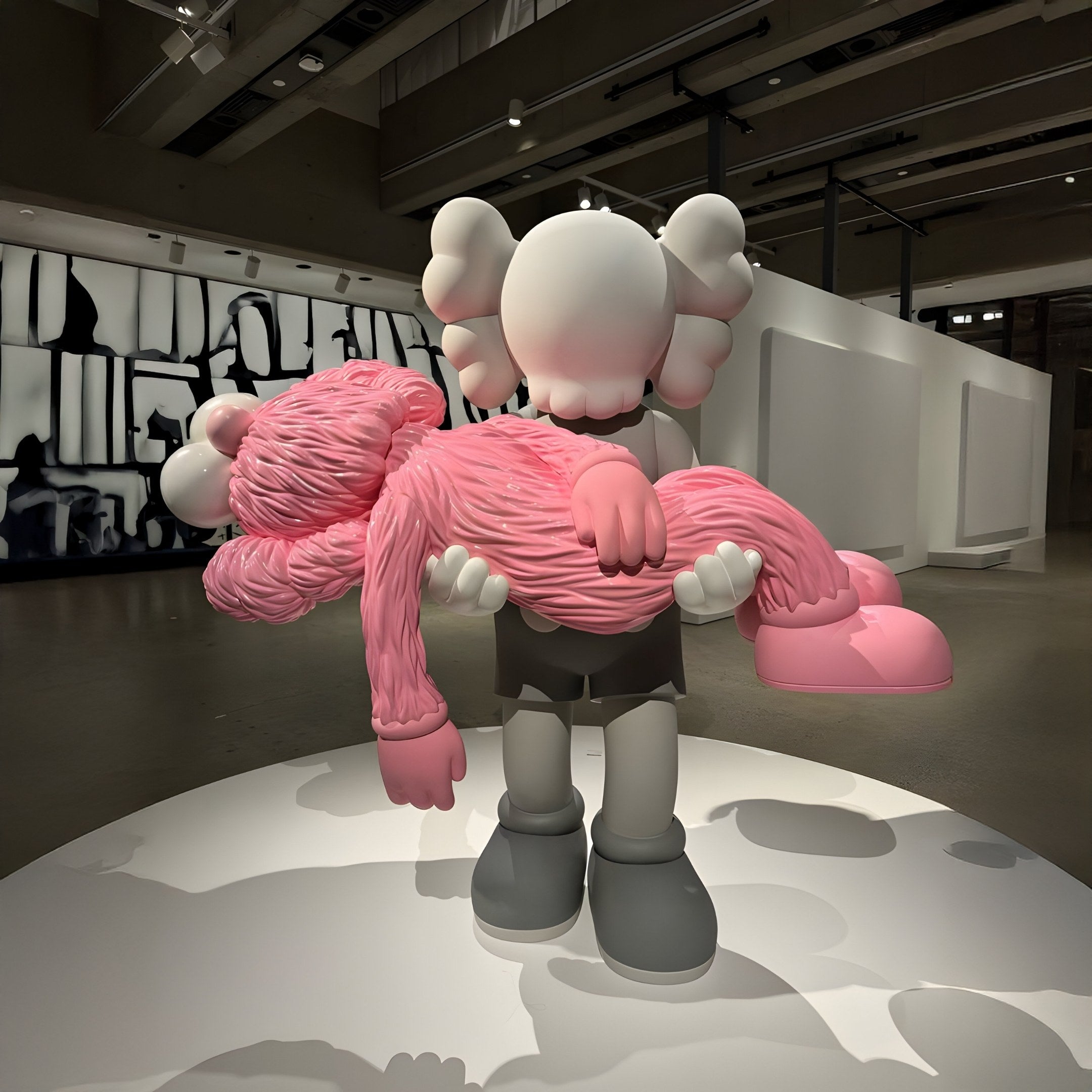 The Giant Sculptures Petal Pink Iconify Rescuing Figure, standing 38cm tall, depicts a cartoon-style character with crossed-out eyes gently cradling a fuzzy pink form. Displayed in a modern gallery with abstract black-and-white art, this piece is both captivating and poignant.