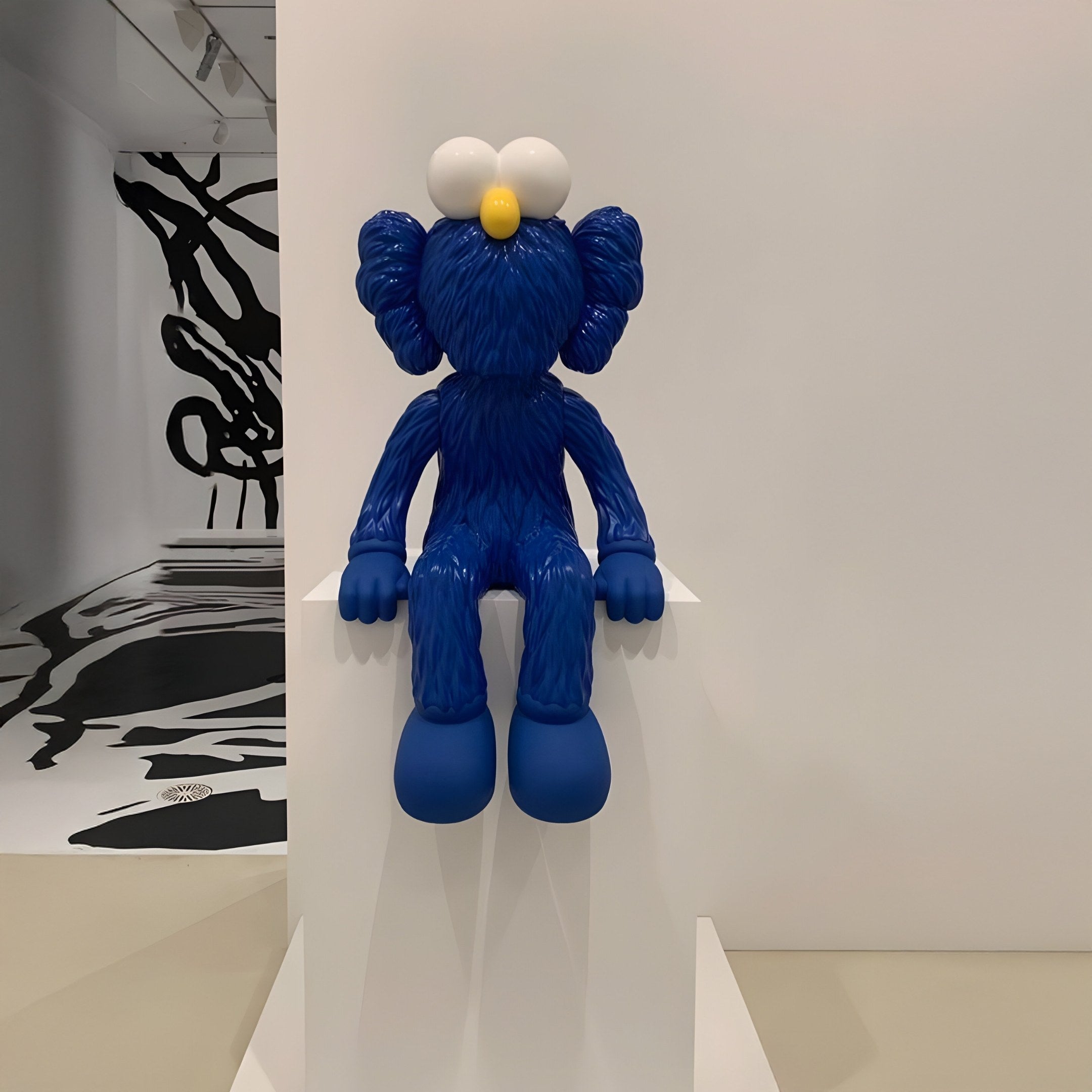 The Deep Blue Iconify Furry Sitting Figure - 40cm by Giant Sculptures, with large white eyes and a yellow nose, sits on a white pedestal. The iconic Pop Art figure lacks facial details, while black abstract designs adorn the gallery walls.