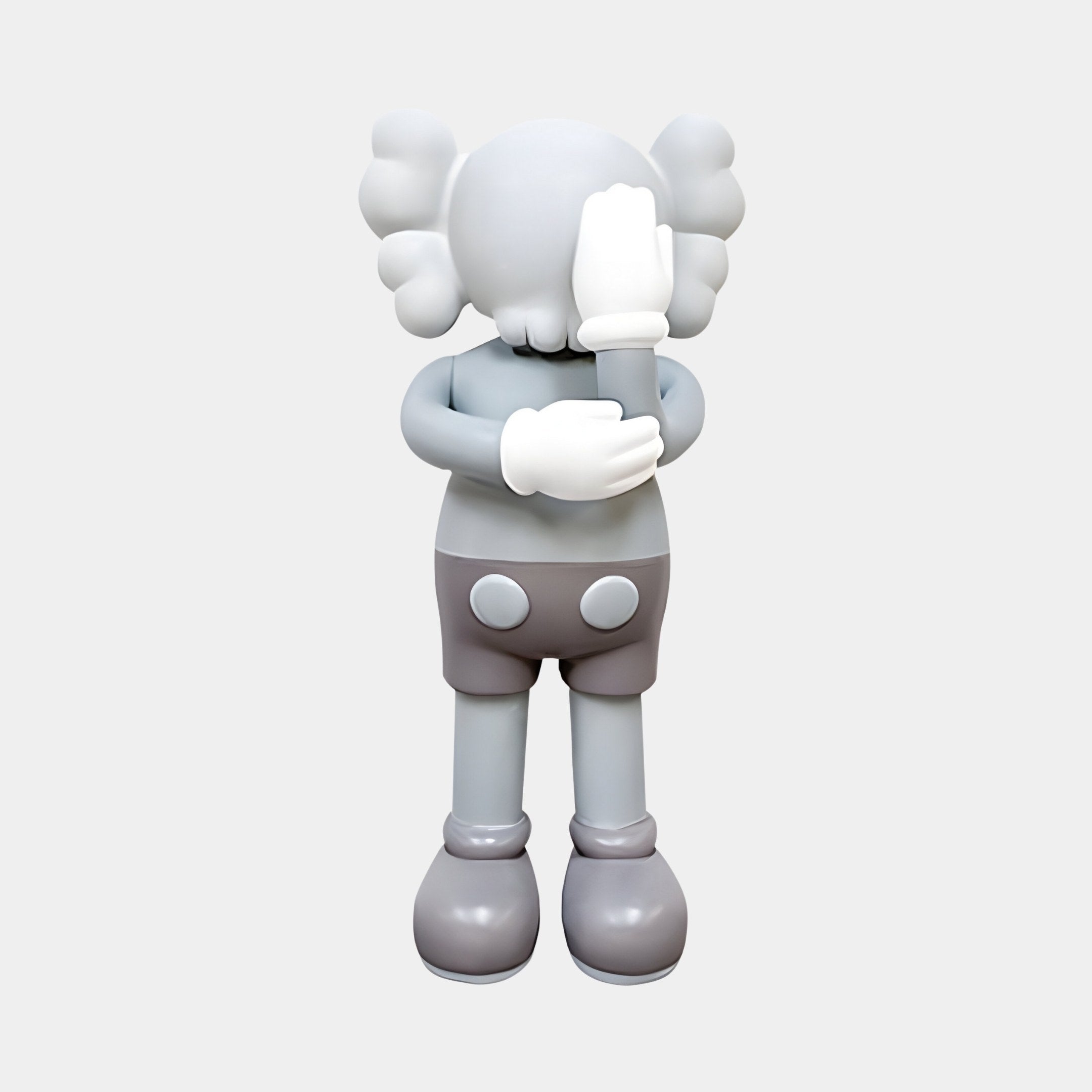 The Soft Grey Iconify IV Dance Sculpture by Giant Sculptures features a cartoon-like figure with a round head, X eyes, large bone-like ears, crossed arms, one gloved hand over its face, wearing shorts and large shoes against a plain backdrop. This contemporary sculpture stands 70cm tall.