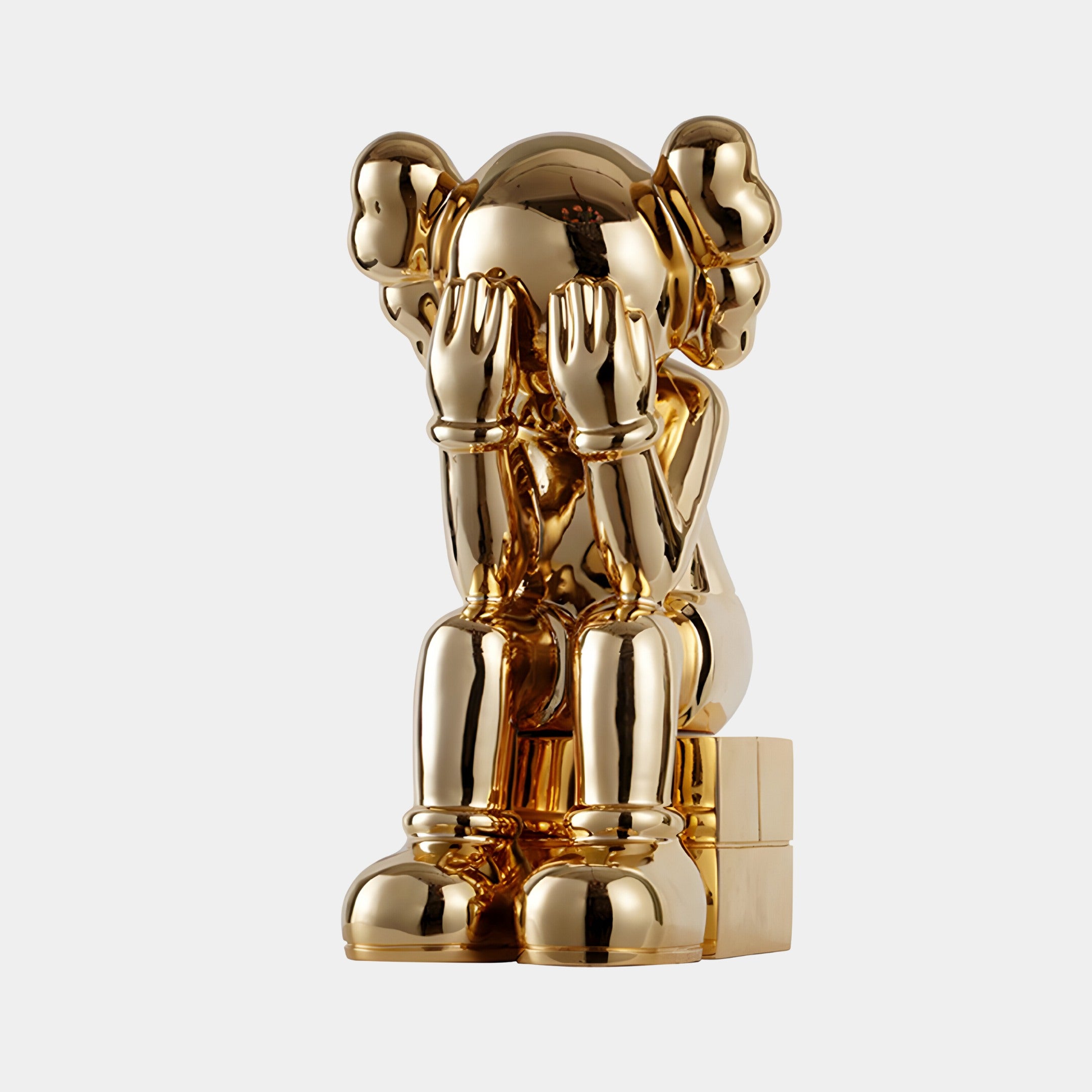 The Antique Gold Iconify Melancholy Sitting Sculpture by Giant Sculptures, measuring 70cm, depicts a figure with cartoonish aesthetics: rounded features and large shoes, sitting with hands covering its face on a small block against a plain background.