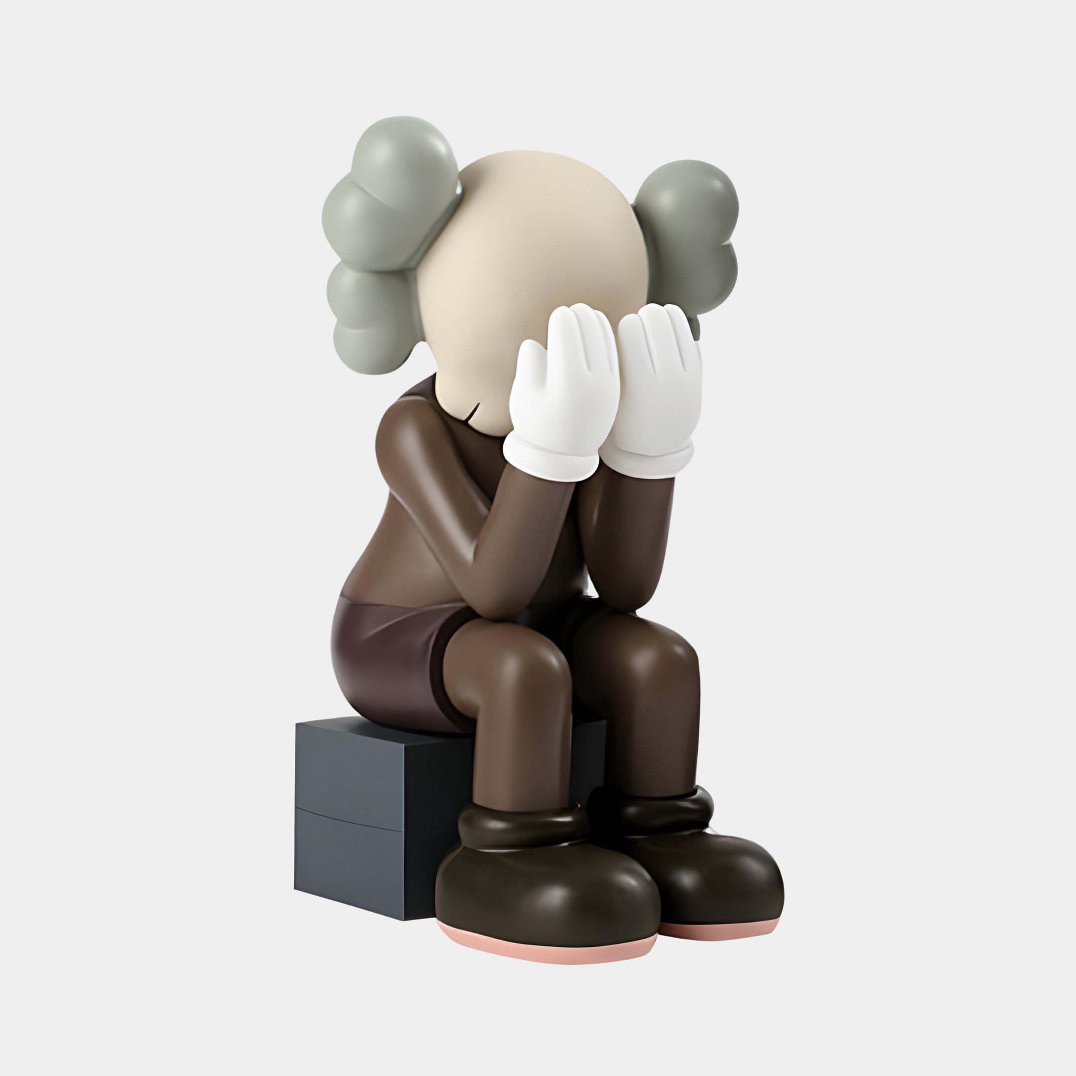 The Hazelnut Brown Iconify Melancholy Sitting Sculpture by Giant Sculptures is a 70cm resin figure with an X-eyed, smooth head, face-covered hands, bulbous gray ears, seated on a block wearing dark clothing and white gloves.