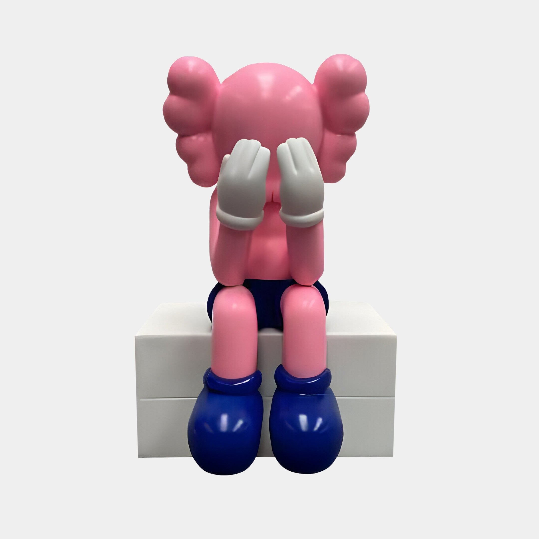 The Petal Pink Iconify Melancholy Sitting Sculpture – 70cm by Giant Sculptures features a figure with gloved hands over its eyes, large round ears, and blue shorts and shoes, sitting on a white block. Its an ideal modern interiors accent.