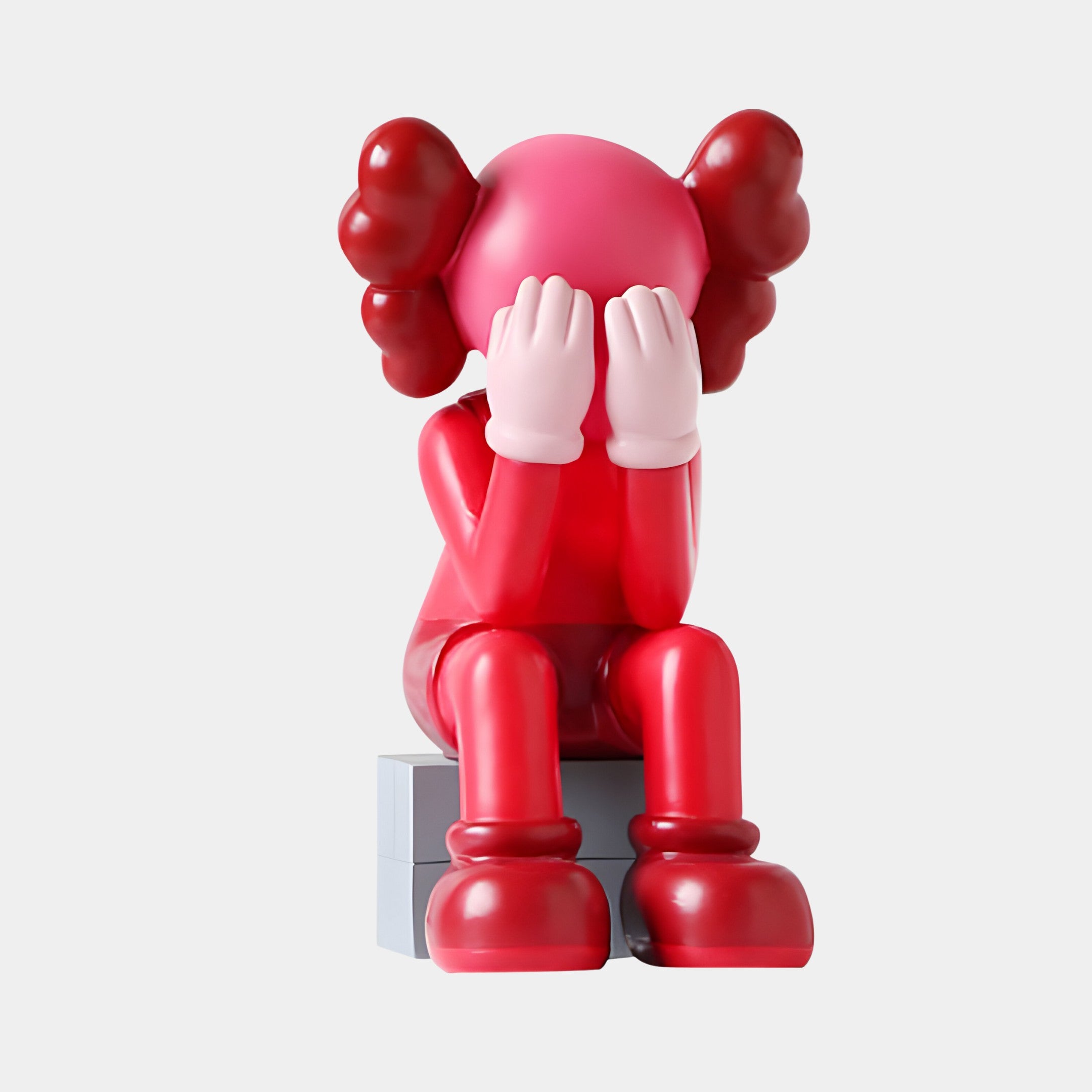 The 70cm Blush Ruby Iconify Melancholy Sitting Sculpture by Giant Sculptures features a red resin figure with cartoon-like rounded ears, sitting on a block and covering its face, blending playful design with introspection.