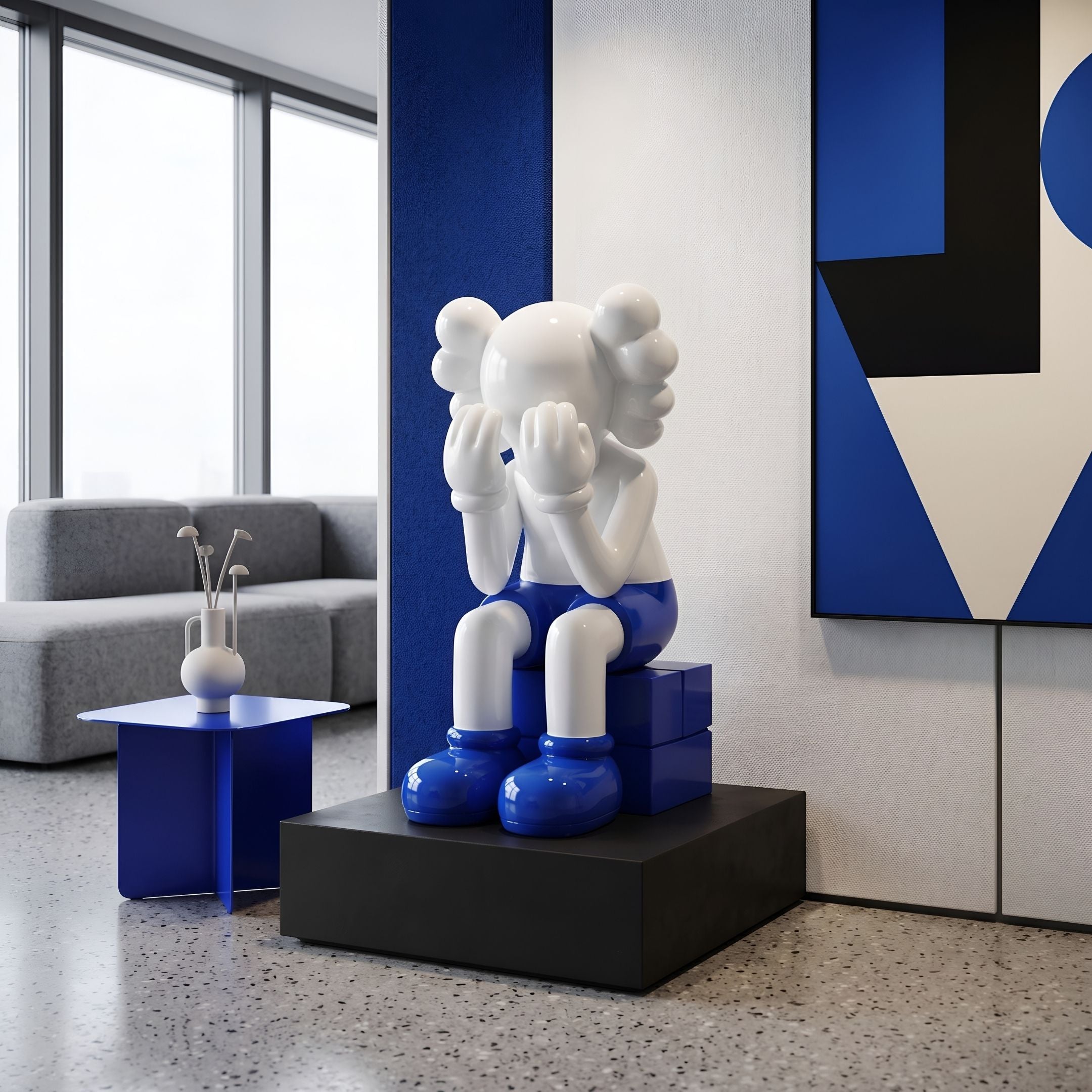 White & Blue Iconify Melancholy Sitting Sculpture, 70cm, seated figure covering face on blue block base, black plinth, in a lounge with grey sofa and blue art.