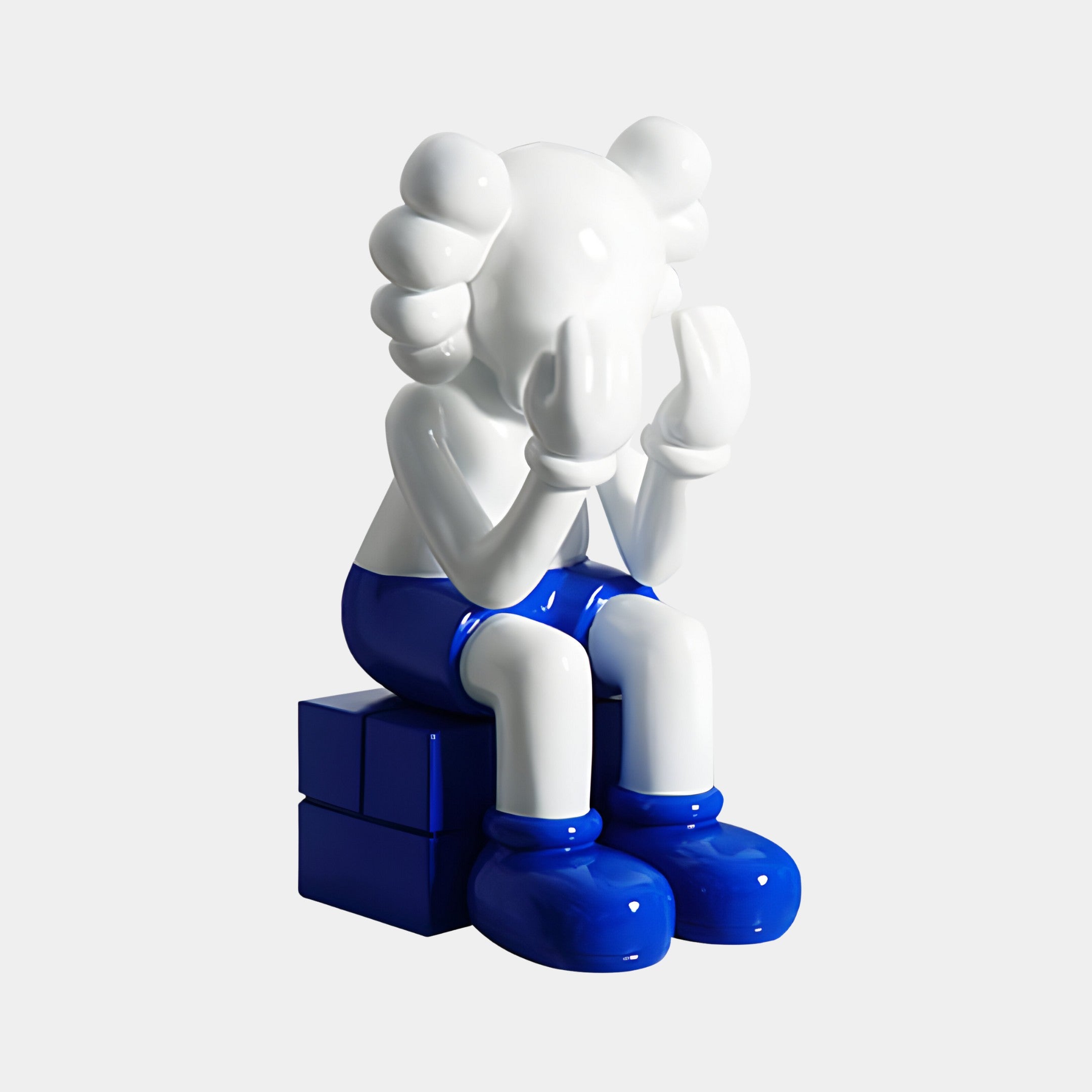 The White & Blue Iconify Melancholy Sitting Sculpture by Giant Sculptures features a 70cm contemporary art piece of a figure covering its face with gloved hands, wearing blue shorts and large shoes while sitting on blue blocks, embodying introspection with its smooth white surface and cartoonish features.