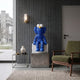 Deep Blue Iconify Furry Sitting Sculpture, 70cm, on a stone pedestal beside a green armchair in a living room with ribbed grey wall paneling.