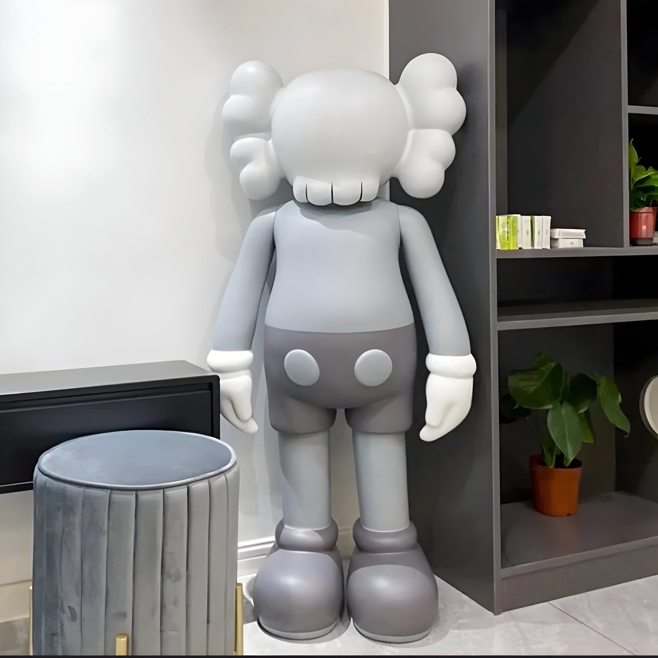 The Soft Grey Iconify Standing Sculpture by Giant Sculptures stands near a black shelf in a room, seamlessly blending with contemporary interiors. The sculpture features X eyes and swirled ears, while nearby are a small plant, books, and a gray upholstered stool.