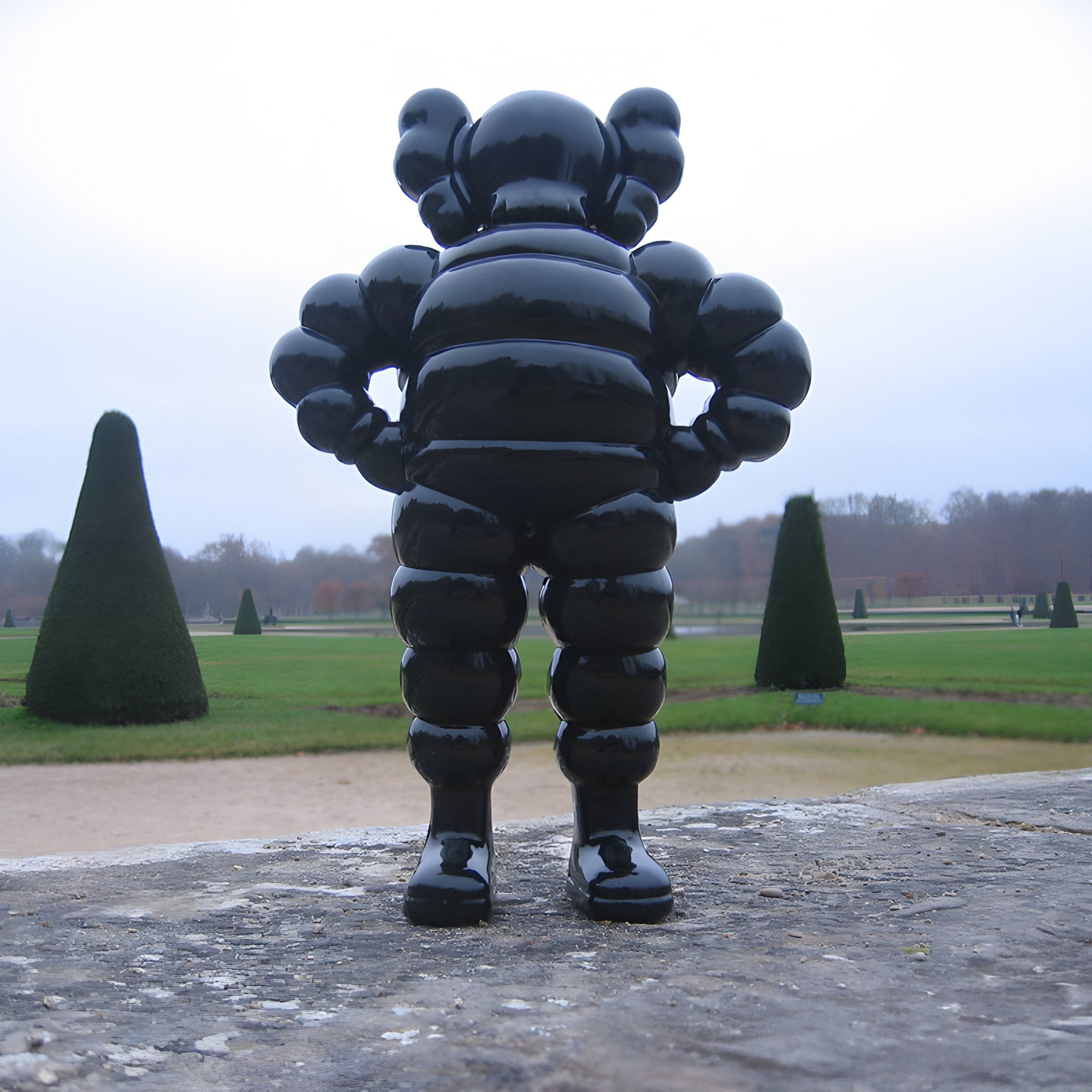 The Midnight Black Iconify Rollo Warrior Sculpture by Giant Sculptures stands 80cm tall on a stone platform, featuring a glossy humanoid figure with distinctive round shapes. Trimmed conical trees and a garden landscape appear under a cloudy sky in the background.