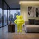 Citrus Yellow Iconify Rollo Warrior Sculpture, 80cm resin figure with hands-on-hips stance, displayed beside a sofa in a modern living room with city view.