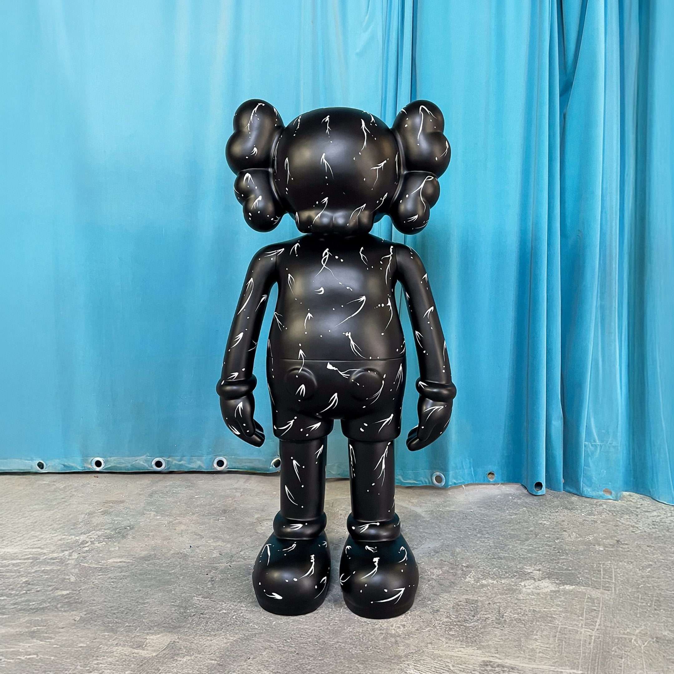 The Black Stone Iconify Standing Sculpture - 130cm by Giant Sculptures features large ears, crossed head stitches, and white markings, standing on a concrete floor before blue curtains—a striking contemporary piece.