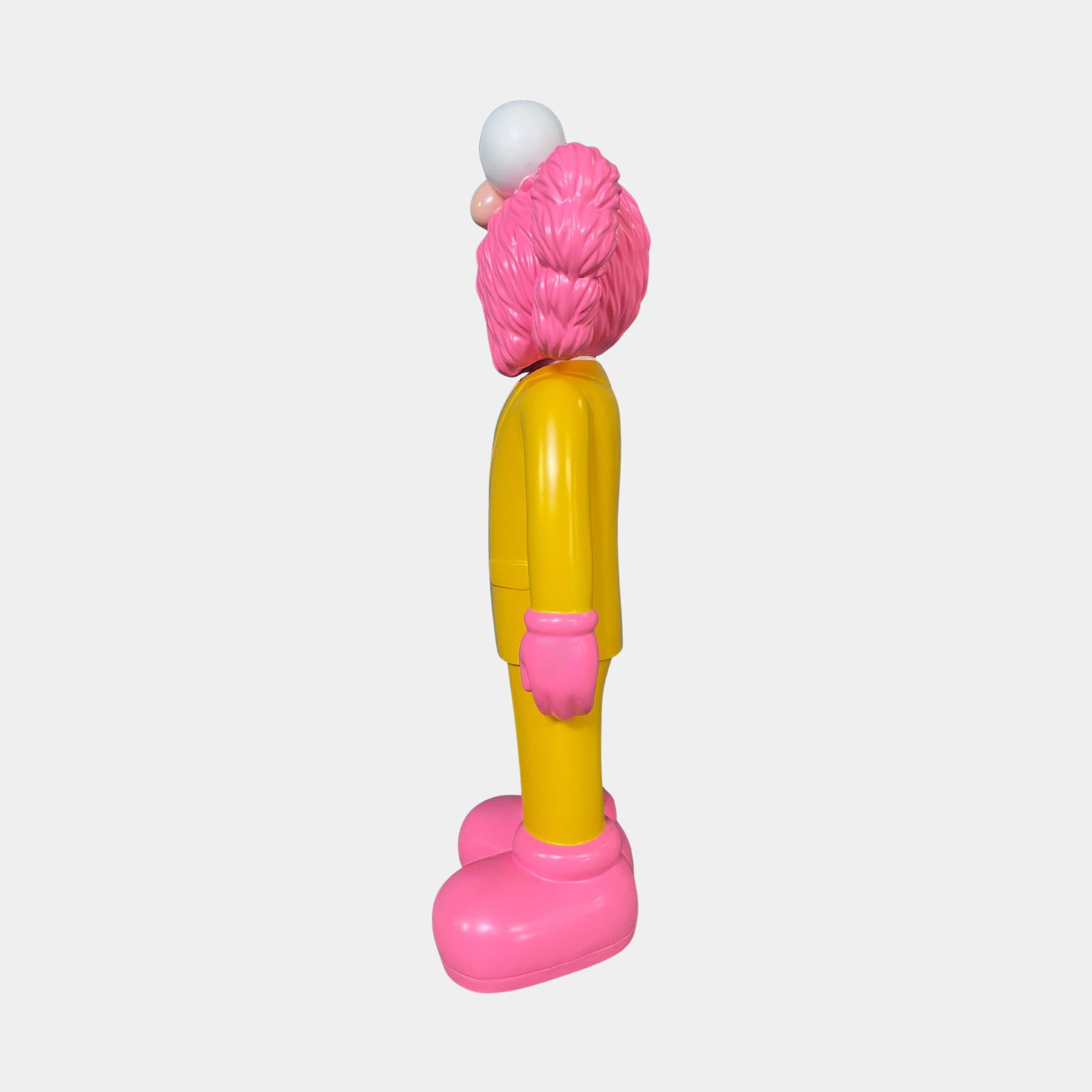 A side view of the Sunbeam Yellow Suit Iconify CEO Sculpture (90cm) by Giant Sculptures, featuring a pink head, large pink gloves and shoes, yellow body, and white round nose—ideal for luxury workspace decor against a plain white background.