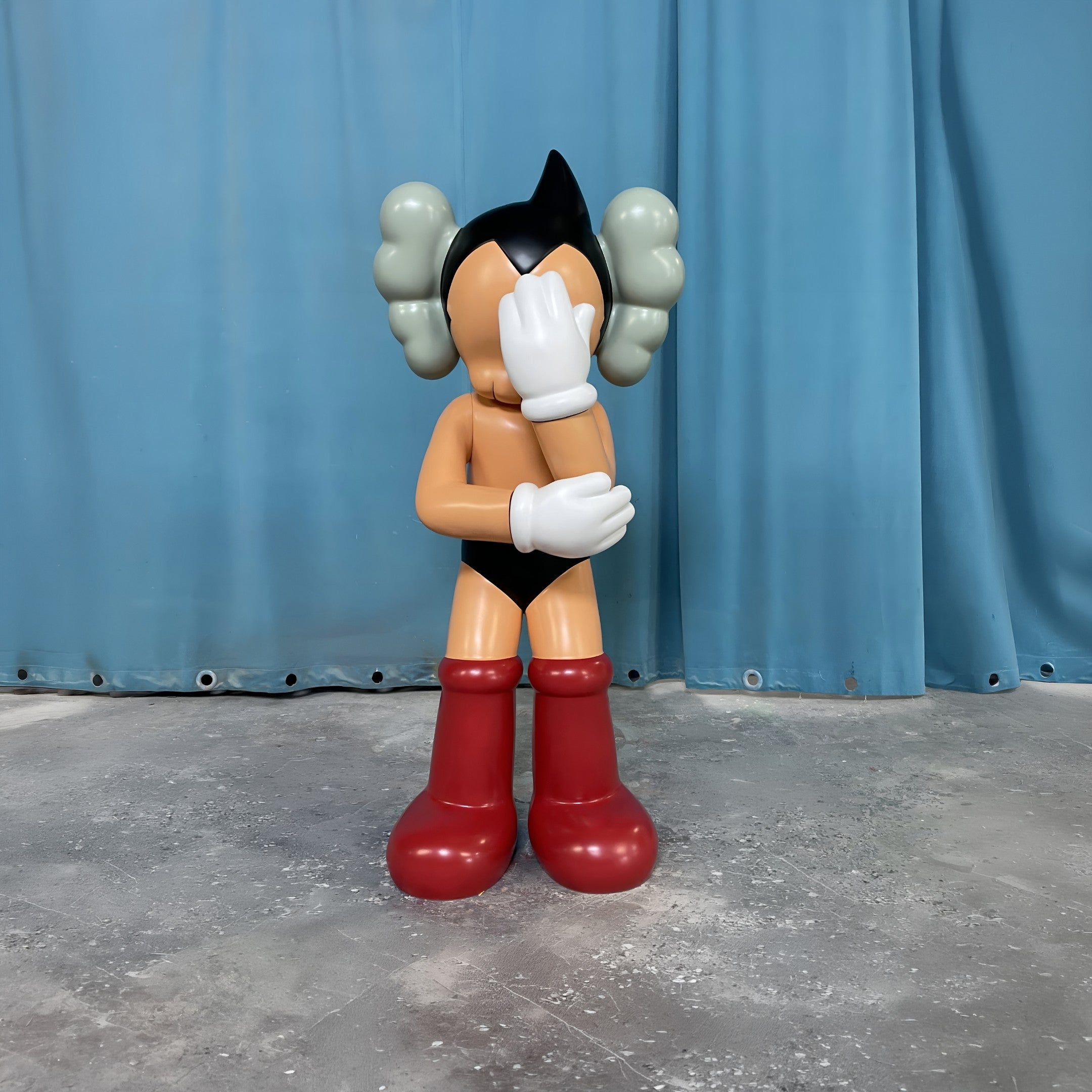 The Giant Sculptures Fiery Red Iconify Cosmic Boy Sculpture - 140cm stands on a concrete floor before blue curtains, covering its face with one hand in a cartoonish gesture of embarrassment or shame.