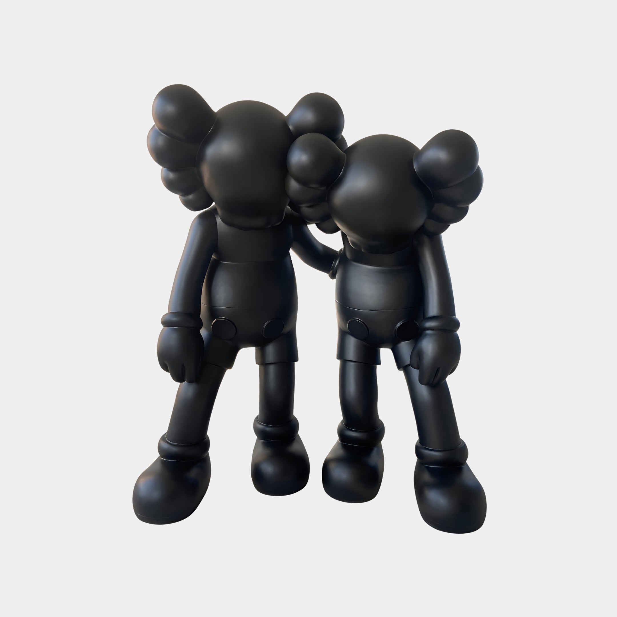 Two Giant Sculptures Midnight Black Iconify Friendship Figures (28cm) with X eyes and gloved hands stand together, one resting its head on the other. This polyvinyl piece makes a bold statement against a plain white backdrop.