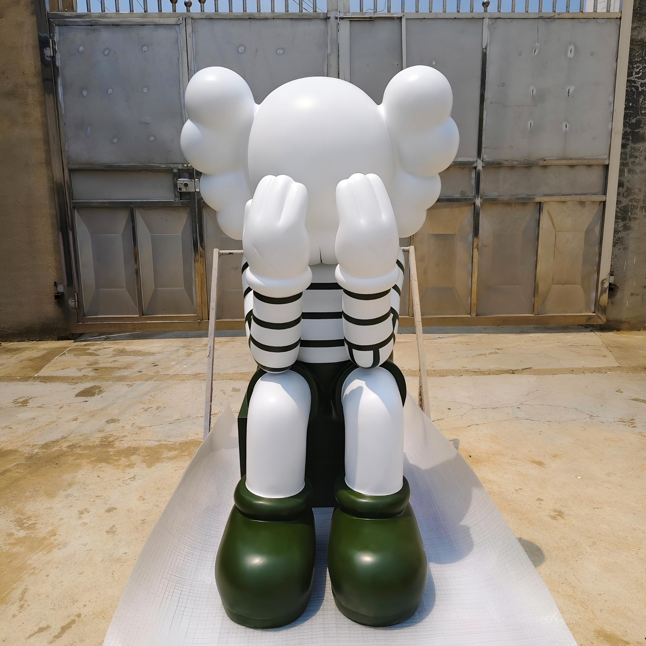 The Giant Sculptures Forest Green Iconify Mime Sitting Sculpture - 135cm photographed in the workshop on a concrete floor against an industrial shutter door, seated with hands to face, striped top and deep forest green boots.
