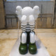 The Giant Sculptures Forest Green Iconify Mime Sitting Sculpture - 135cm photographed in the workshop on a concrete floor against an industrial shutter door, seated with hands to face, striped top and deep forest green boots.
