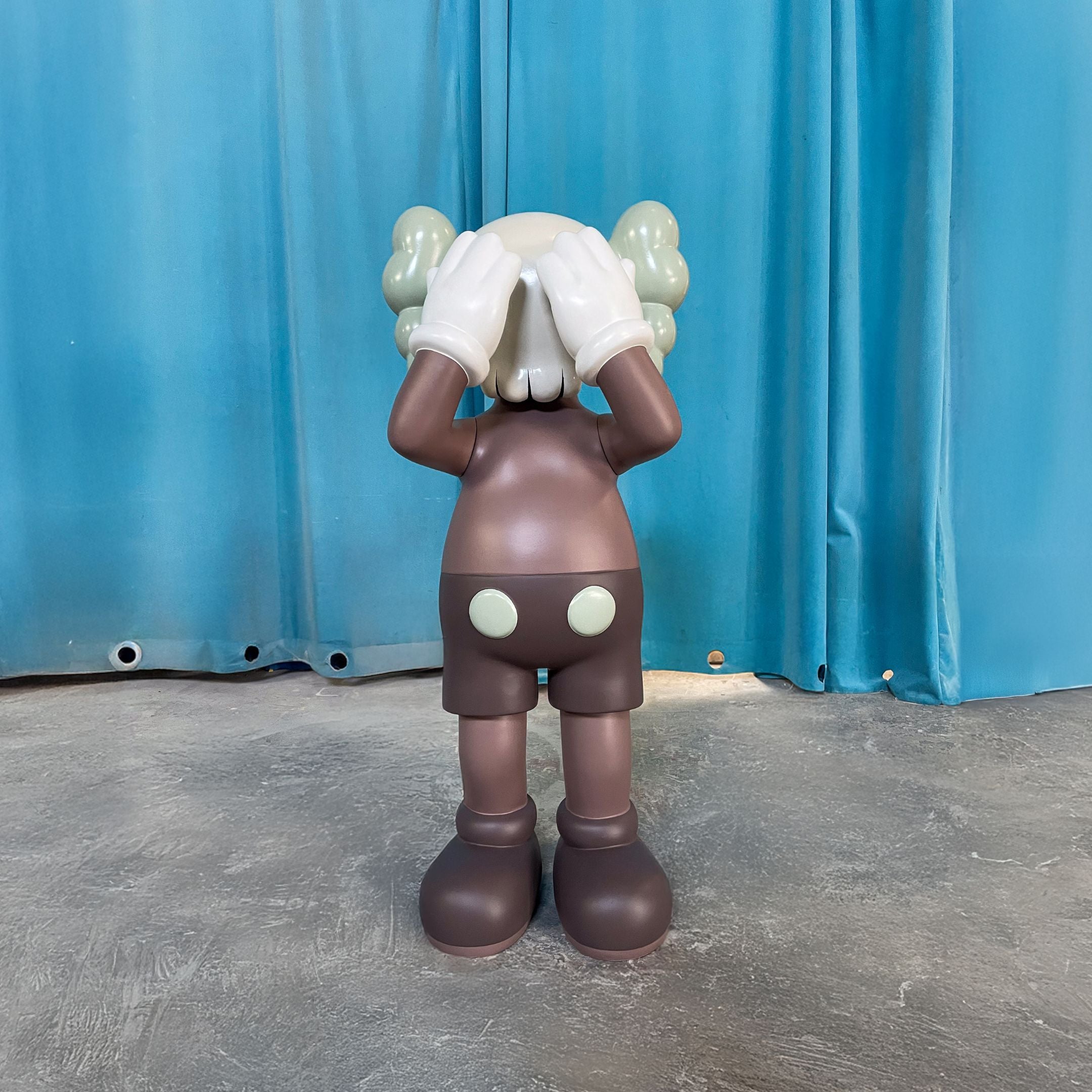 The Hazelnut Brown Iconify Silent Cry Sculpture (91cm) by Giant Sculptures stands on a gray floor before blue curtains, covering its face with gloved hands—a whimsical touch of contemporary art decor.