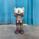 The Hazelnut Brown Iconify Silent Cry Sculpture (91cm) by Giant Sculptures stands on a gray floor before blue curtains, covering its face with gloved hands—a whimsical touch of contemporary art decor.