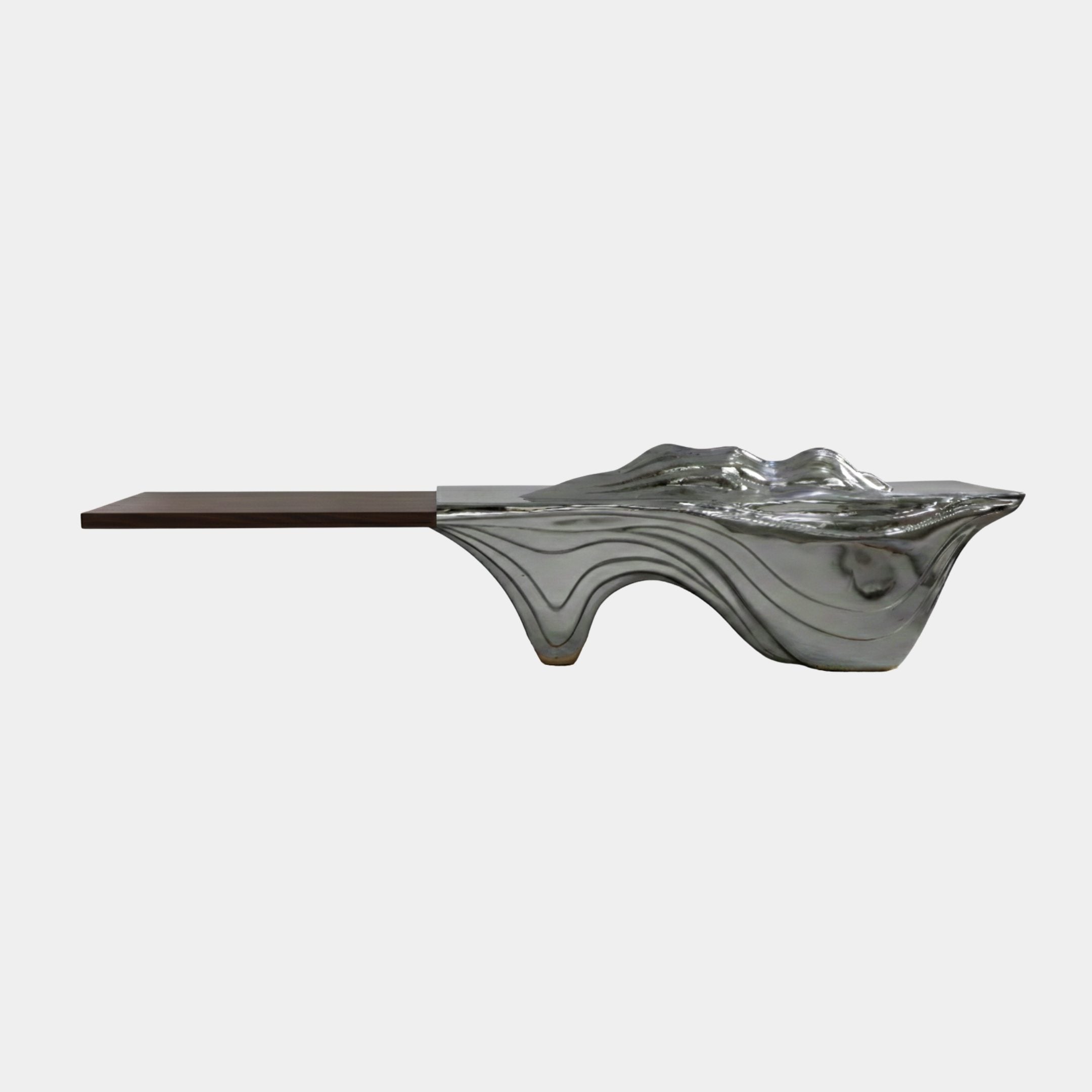 Polished chrome Fluid Wave Lobby Console by Giant Sculptures, its mirror-bright fibreglass form flowing into a long dark wood plank, shown side-on against white.