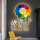 The Clovelle Floral Bliss UV Wood Carving LED Wall Art by Giant Sculptures showcases two figures with faces and bodies of colorful flowers, surrounded by rainbow-like petals. Below, a yellow chair complements the vibrant floral motif beautifully.