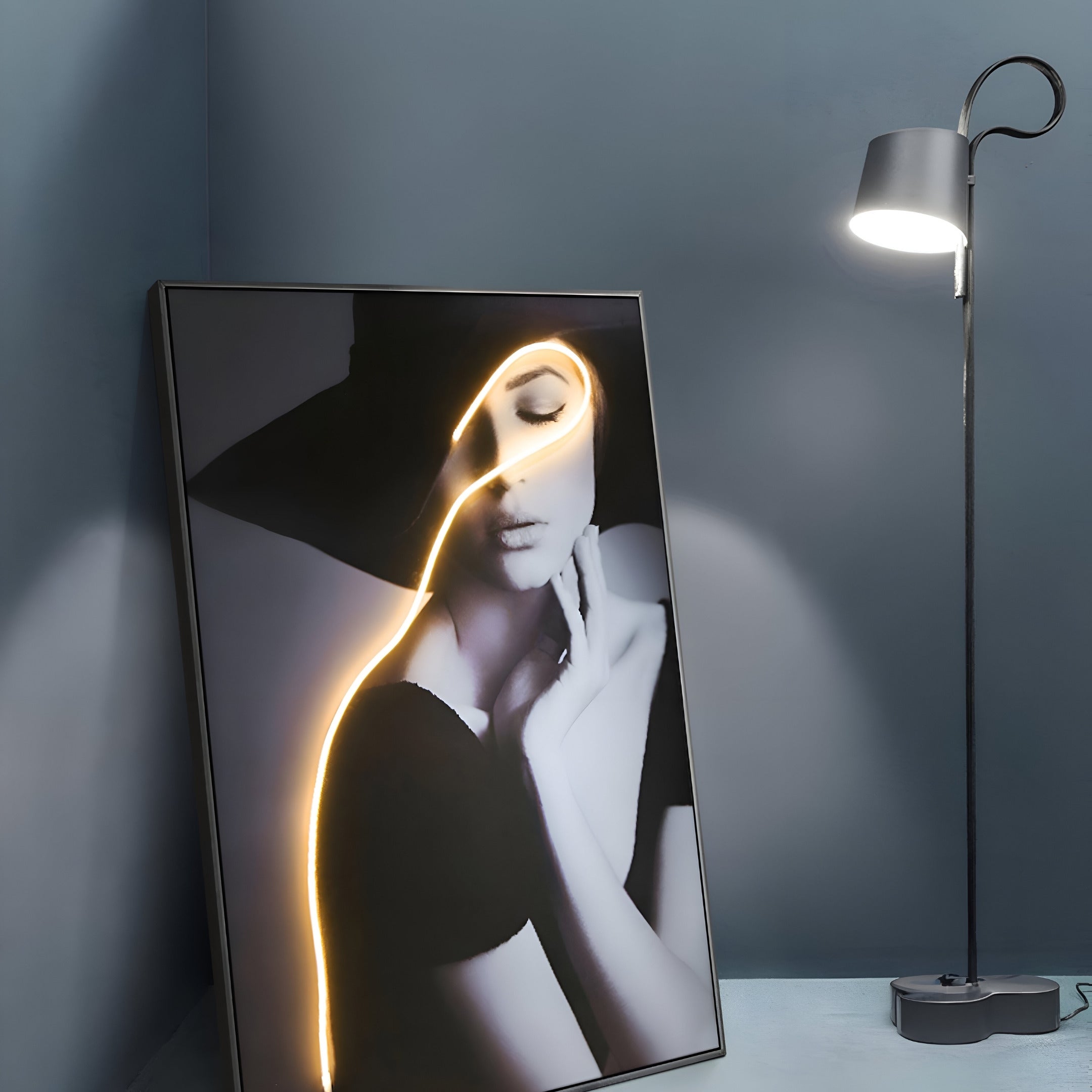 The Sylva Noir Muse Canvas LED Wall Art by Giant Sculptures features a modern black and white portrait of a woman in a large hat against a blue wall. An artistic light strip adds elegance, while a contemporary floor lamp softly enhances its allure.