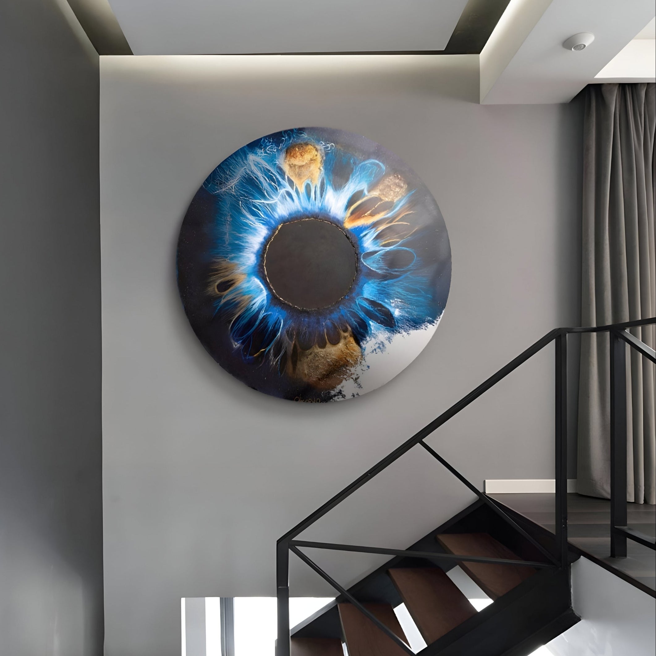 A modern staircase with wooden steps and a black railing ascends, and on the gray wall, the Genesis Earth Tones 3D Resin Crystal Porcelain LED Wall Art by Giant Sculptures displays swirling blue, black, and gold patterns resembling an abstract eye. Earth tones blend beautifully as natural light partially illuminates the space.