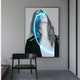 The Elysian Veiled Grace 3D LED Wall Art by Giant Sculptures features a framed black-and-white photo of a long-haired person, elegantly illuminated with a striking neon blue curve. It adorns a gray wall next to a black chair and wooden surface, exuding contemporary aesthetics.