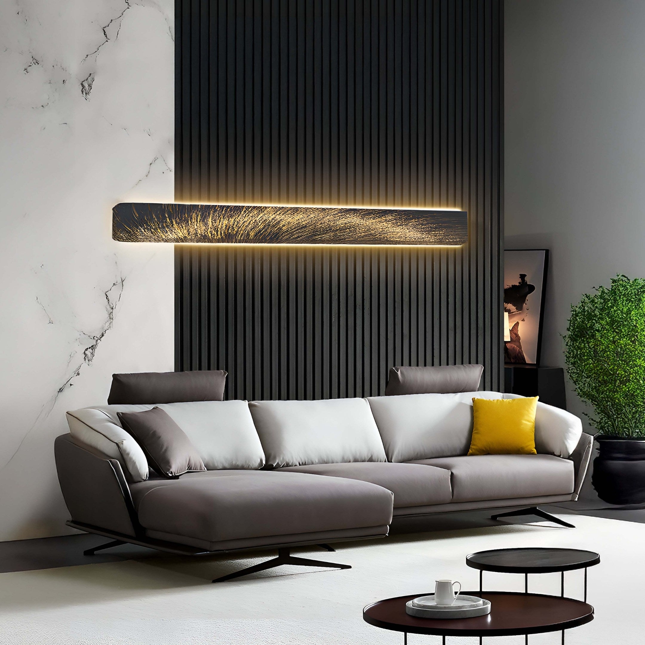 In a modern living room, a gray sectional sofa with a yellow cushion faces two round coffee tables. Behind it stands Giant Sculptures Radiance Golden Canvas LED Wall Art on a black panel. To the left is a marble wall, and to the right is a potted plant.