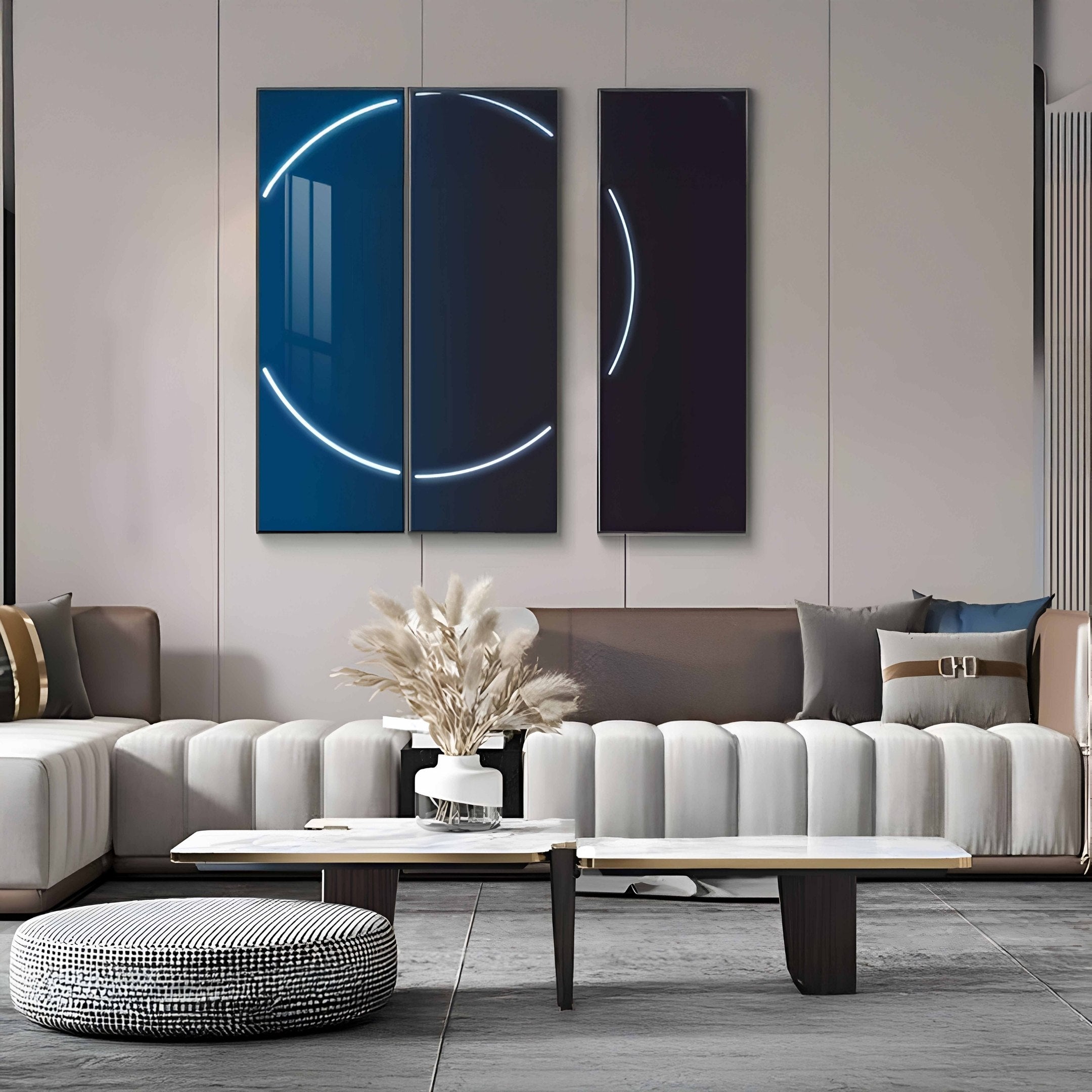 A modern living room showcases a beige sofa with decorative pillows, a round pouf, and a sleek coffee table. On it sits the Helios Cobalt Crystal Porcelain Vase with pampas grass. LED wall art by Giant Sculptures, featuring abstract neon lines on dark panels, completes the minimalist aesthetic.