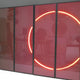 The Helios Ruby Crystal Porcelain LED Wall Art by Giant Sculptures features four glass panels tinted with ruby crystal and two central panels displaying glowing red lines in a partial circle, resembling LED wall art, and reflecting a blurred indoor setting.