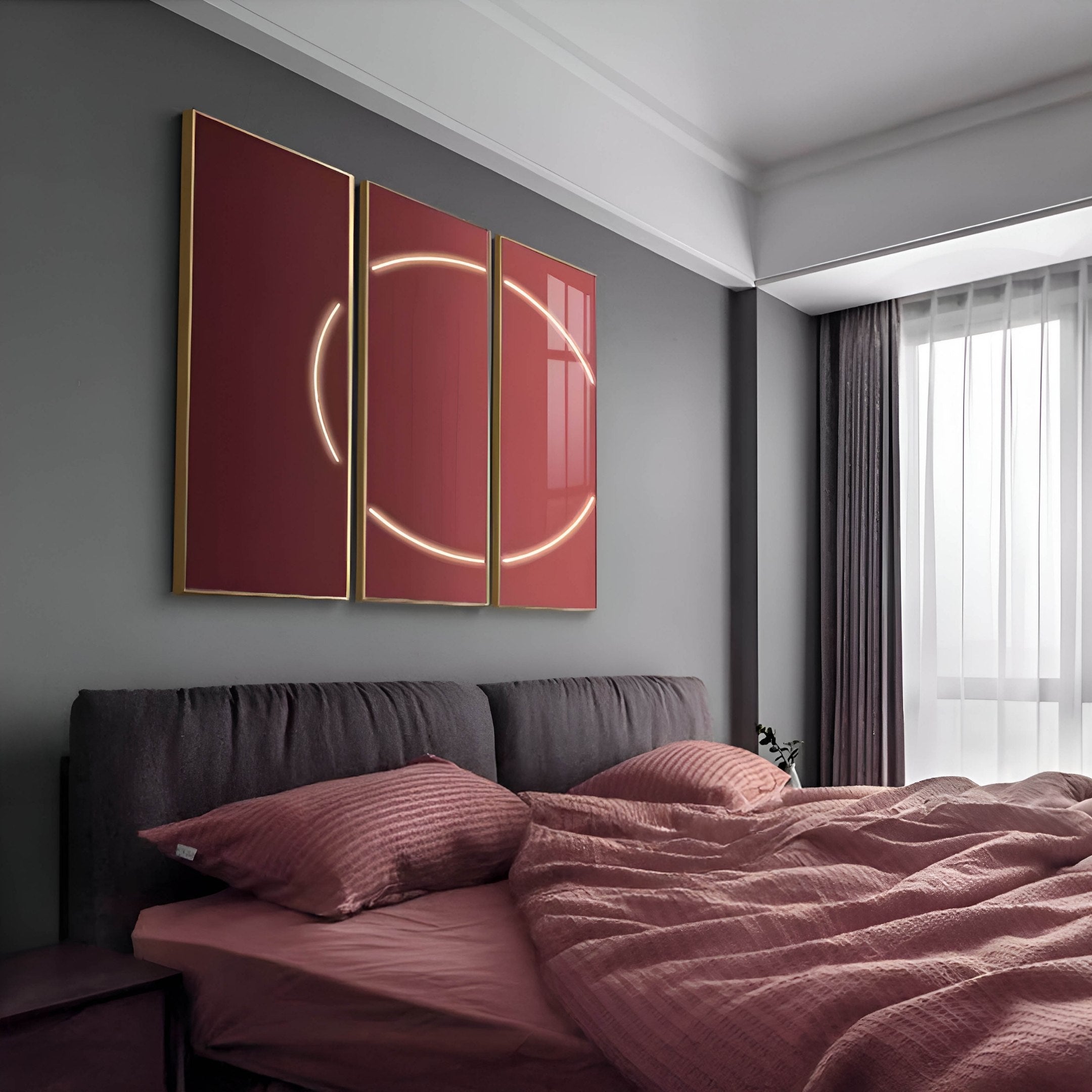 A modern bedroom includes a gray headboard with pink bedding. Above the bed, three vertical panels showcase the Giant Sculptures Helios Ruby Crystal Porcelain LED Wall Art featuring a glowing circle design. Natural light floods in through a large window with sheer curtains.