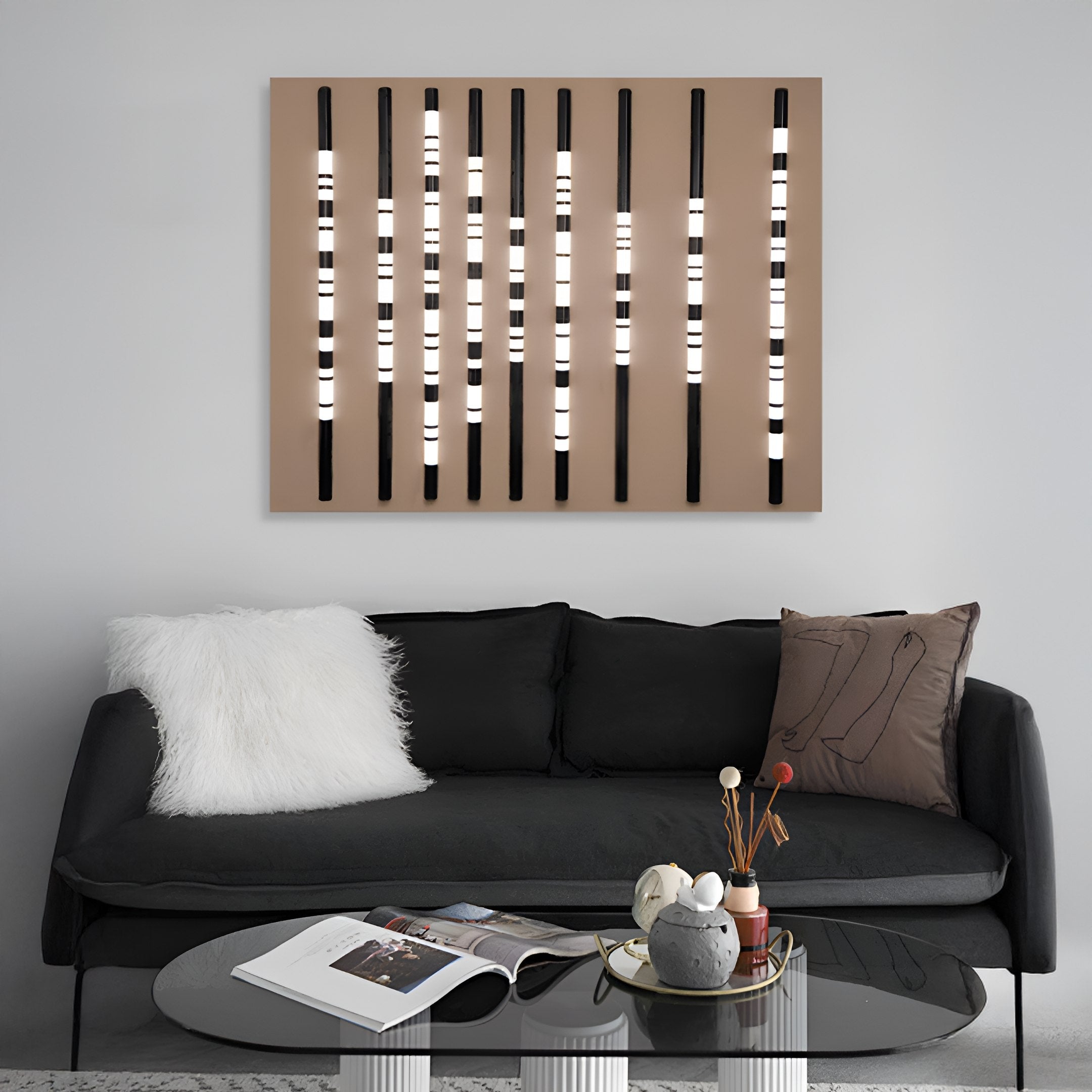 A modern living room includes a black couch with a white fluffy pillow and a brown abstract art pillow. A round glass table holds a magazine and decor. The Strata Taupe LED Wall Art by Giant Sculptures adds vibrancy to the light gray wall, ideal for modern homes.