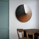 In a contemporary space, the Orbit Copper Hand-Painted Canvas 3D LED Wall Art by Giant Sculptures featuring black, brown, and beige shades adorns the wall. Below it rests a wooden table with two curved-back chairs. On the left, a muted green partition is partially visible.