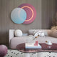 A modern living room features a beige sofa with pastel cushions and a glass-top table. On the wall is the Giant Sculptures Lunar Blush Sky Crystal Porcelain 3D Yellow LED Wall Art with glowing circles. A sculpture, candle, and coffee cup adorn the table along with a round ottoman and rug in Lunar Blush Sky tones.