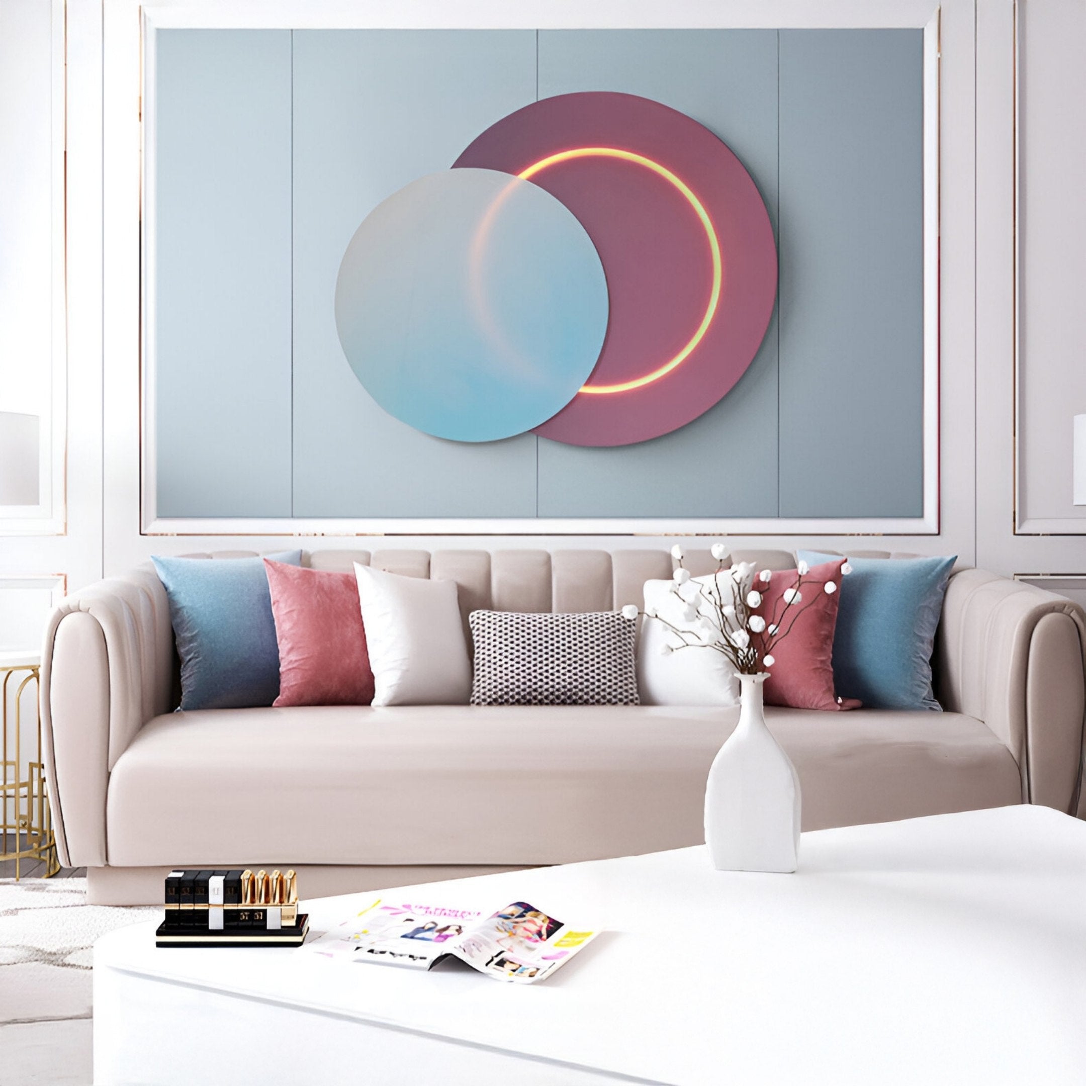 A modern living room features a beige sofa with pink, blue, and white cushions. Above it hangs the Lunar Blush Sky Crystal Porcelain 3D Yellow LED Wall Art by Giant Sculptures, showcasing intersecting circular shapes. A white coffee table with magazines and a vase of branches completes the serene scene.