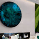 A modern interior features a striking large circular blue and green abstract artwork. Below, sleek white chairs and tables are complemented by the Lumea Textured Turquoise 3D LED Crystal Porcelain Wall Art by Giant Sculptures. A green textured wall art piece is visible on the right.