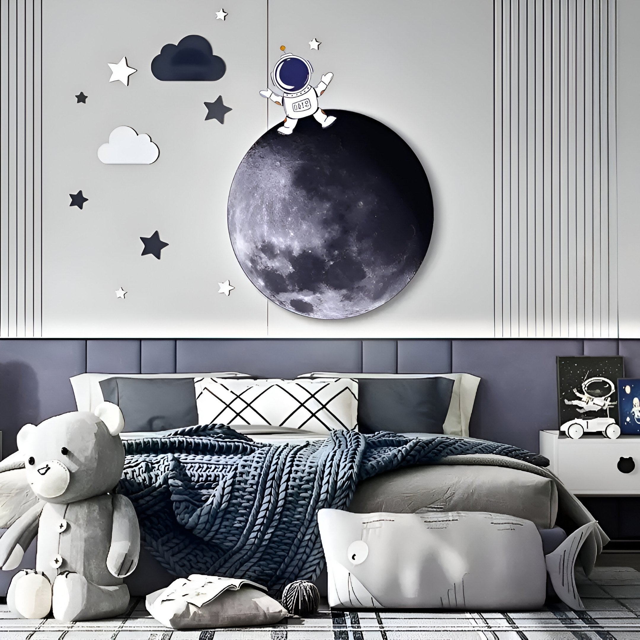 A childrens bedroom is adorned with the Cosmos Astronaut Dream UV Wood Carving LED Wall Art by Giant Sculptures, featuring an astronaut on a moon, stars, and clouds. The bed has a gray bedspread with a teddy bear and whale pillow, complemented by a light gray headboard and striped wallpaper.