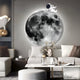 A modern living room features a large moon wall mural and the captivating Cosmos Astronaut Drift LED Wall Art by Giant Sculptures. It includes a gray couch, white pillows, round ottoman, decorative plants, and a cloud-shaped white wall lamp.