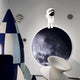 A playful space-themed room features a moon mural, a blue and white rocket model, and Giant Sculptures Cosmos Astronaut Reflection UV Wood Carving LED Wall Art. A modern white chair sits in the foreground with round wall decorations resembling planets surrounding it.