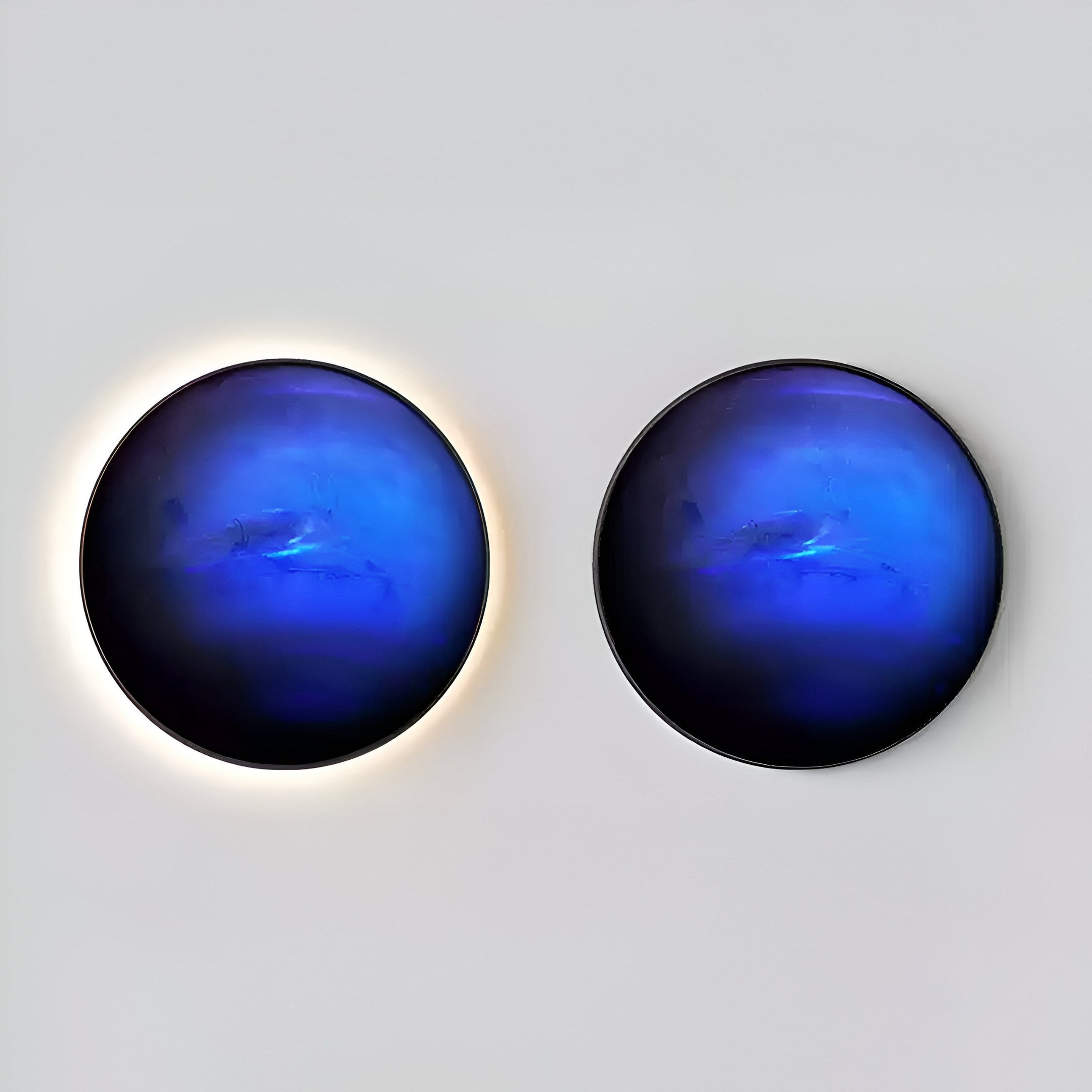 Two circular blue wall lights on a plain background evoke the look of Galaxia Neptune Crystal Porcelain LED Wall Art by Giant Sculptures. The left light has a glowing halo, while the right lacks it. The blue hue features subtle dark and light variations, resembling the night sky from Galaxia Neptune.