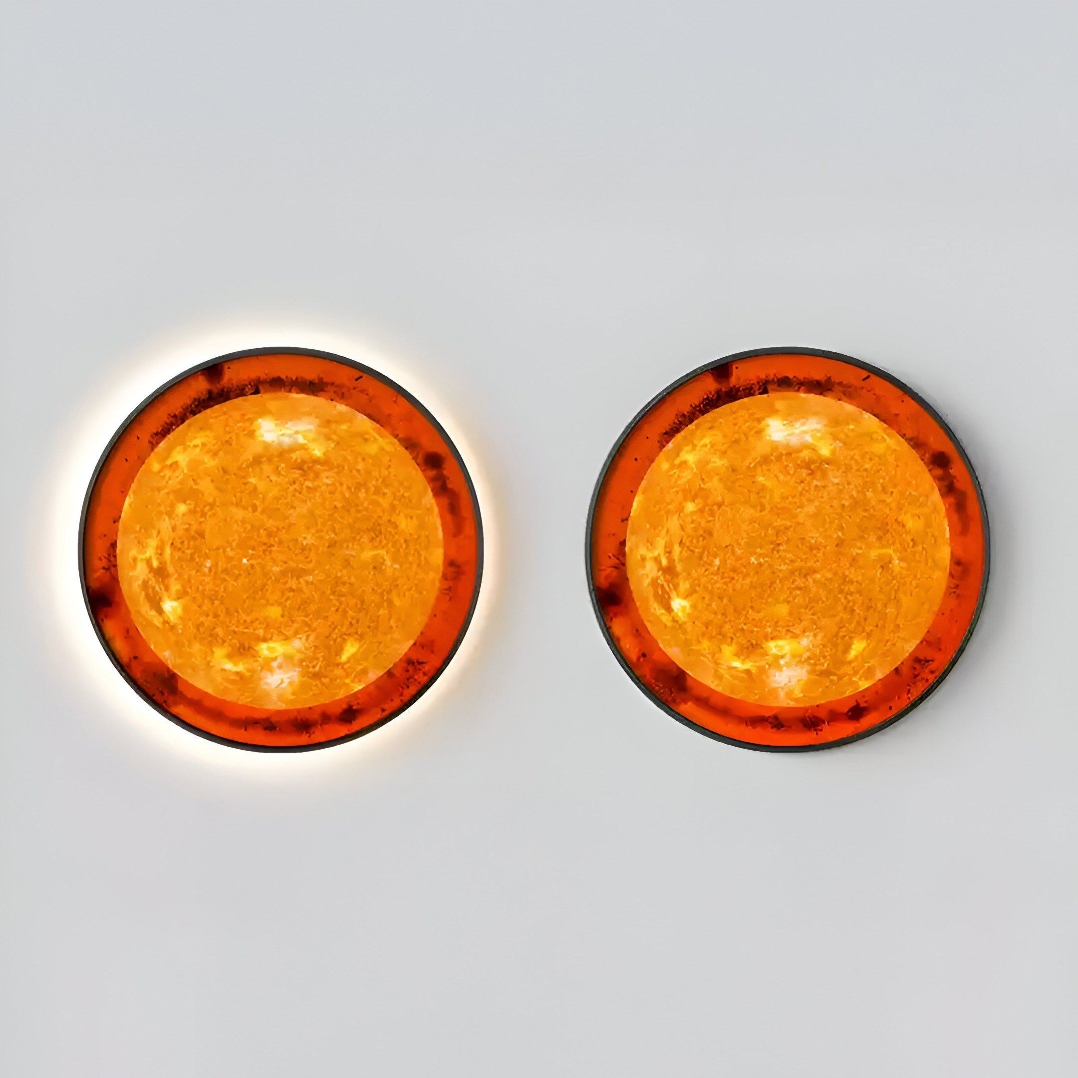 The Galaxia Sun Crystal Porcelain LED Wall Art by Giant Sculptures features two amber-hued wall lights with dark rims on a light gray backdrop. Their textured surfaces create an artistic sunburst effect, with the left fixture glowing subtly within its brushed aluminum frame.