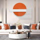 A modern living room features a white sofa adorned with orange, white, and black cushions. A round marble coffee table displays books and a plant. Above the sofa hangs Giant Sculptures Aeon Sunset Ember Leather 3D LED Wall Art, showcasing geometric shapes in orange, white, and gray.