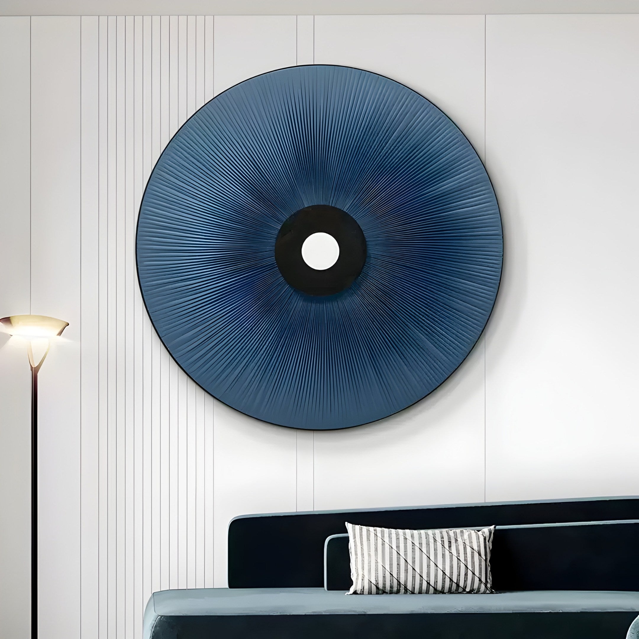 The enticing Vanguard Midnight Indigo Leather 3D LED Wall Art by Giant Sculptures, showcasing an indigo hue and black center with a white dot, adorns the white wall. Below, a dark green couch with a striped pillow is visible as a floor lamp on the left casts light on this artwork.