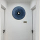 A minimalist hallway with white walls and doors features a blue circular wall art piece with an indigo hue. The black center adds modern elegance, echoing the aesthetic of Giant Sculptures Vanguard Midnight Indigo Leather 3D LED Wall Art.