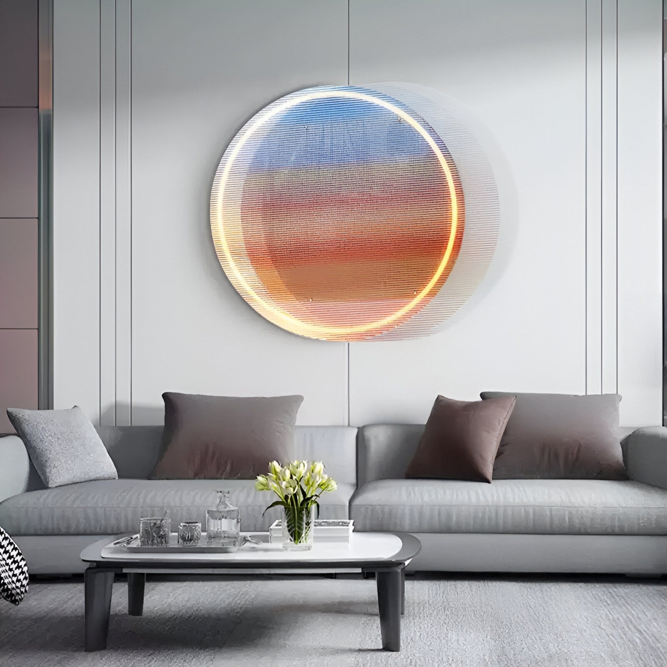 A modern living room showcases a gray sofa with assorted pillows. A white coffee table with a vase of flowers sits in front, while behind the sofa, Giant Sculptures Chromia Sunrise Glow Acrylic LED Wall Art illuminates the space with its circular abstract design in warm and cool tones.