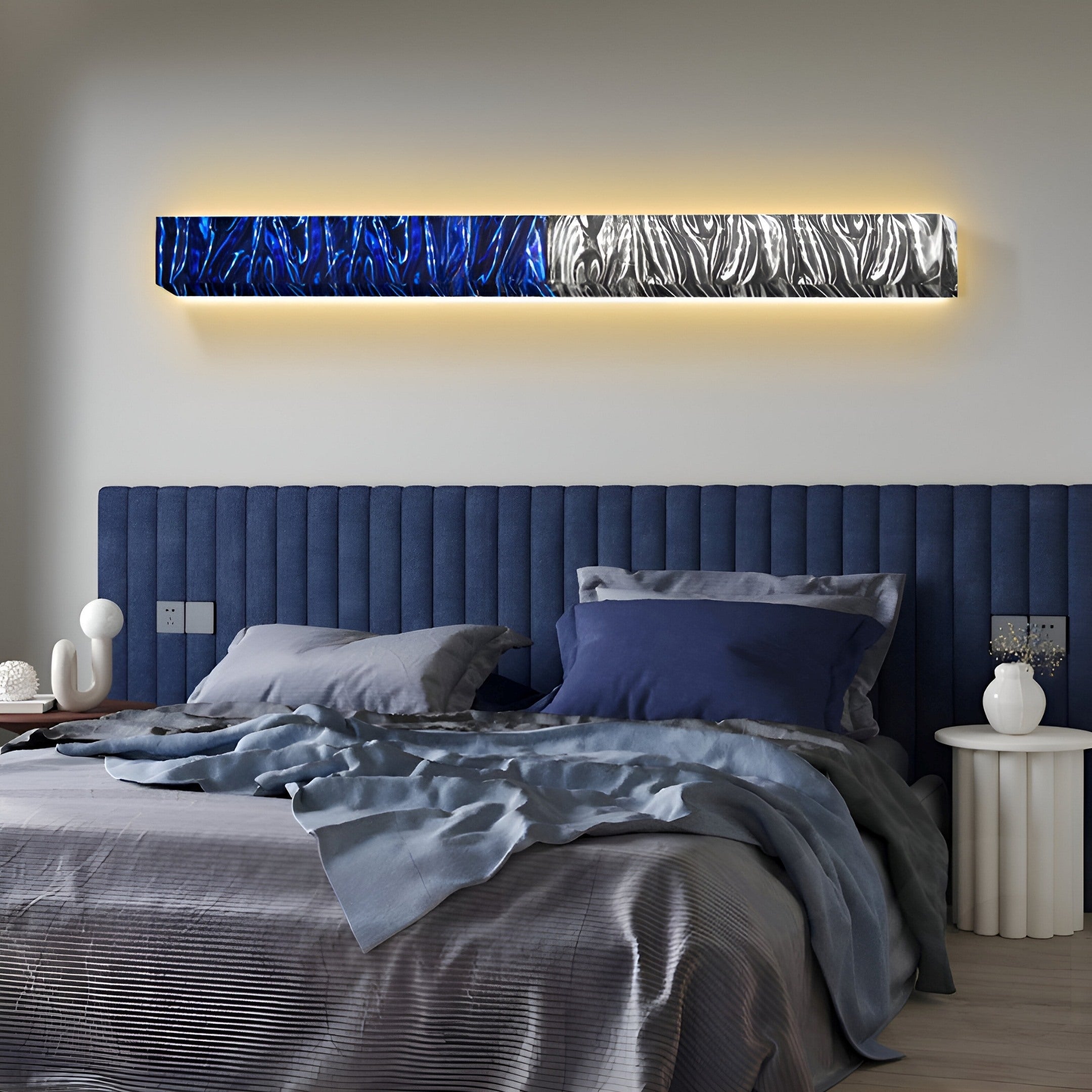 Modern elegance defines this bedroom with a dark blue padded headboard and gray bedding. Above the bed, Giant Sculptures Textura 3D Blue and Silver Leather Design Wall Art enhances the space. A white side table holds decorative items, adding to the sleek aesthetic with leather accents.